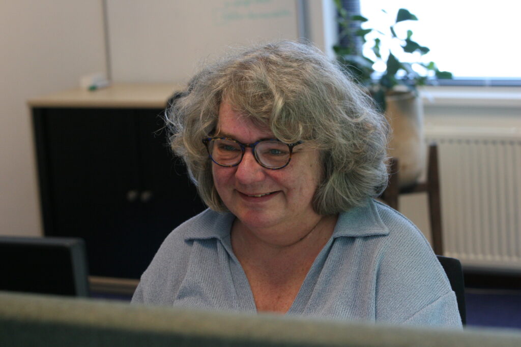 Een vrouw (onze collega José) zit lachend achter haar bureau.